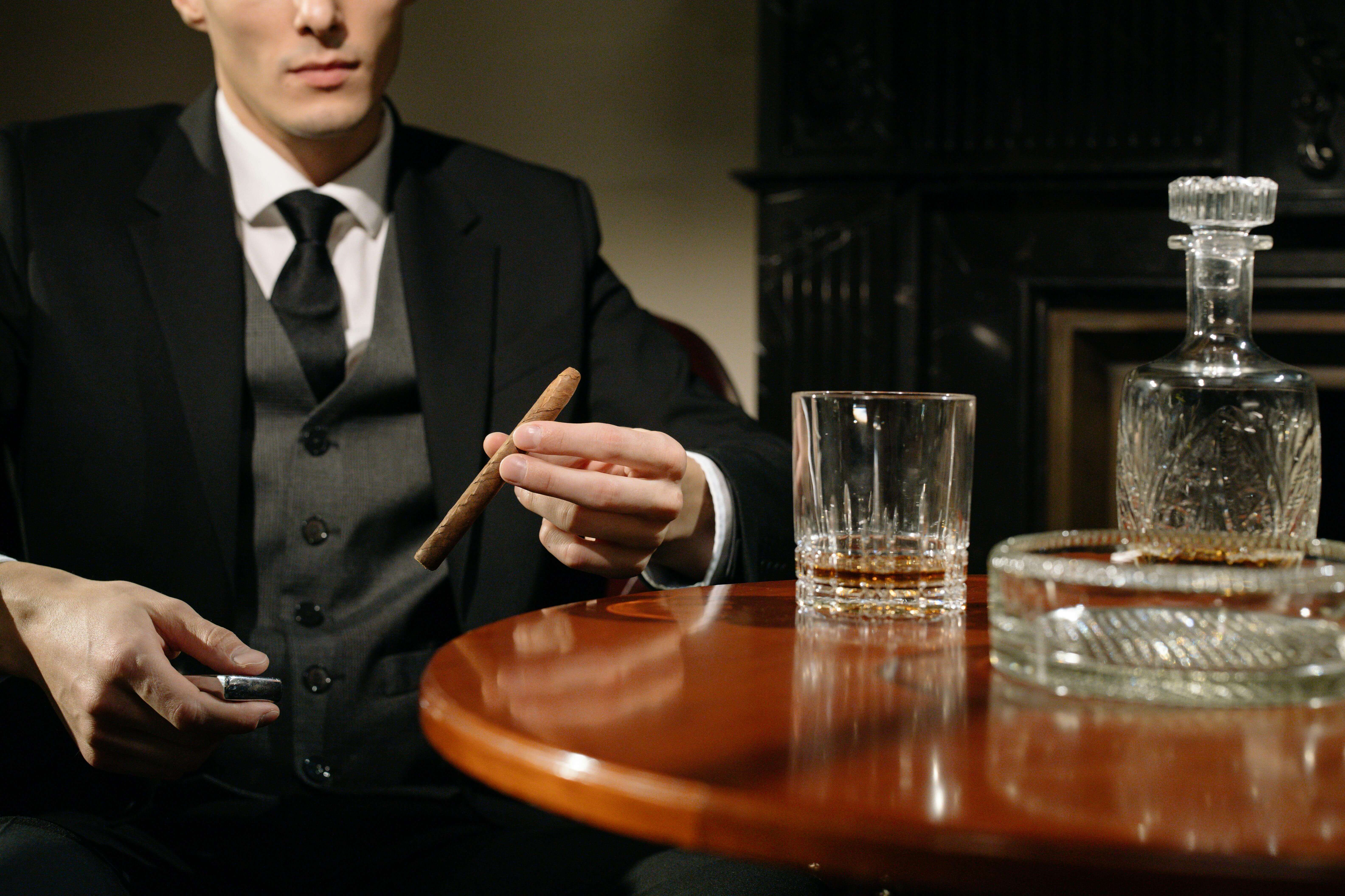 a man in a suit and tie holding a cigar at a bar table with a drink